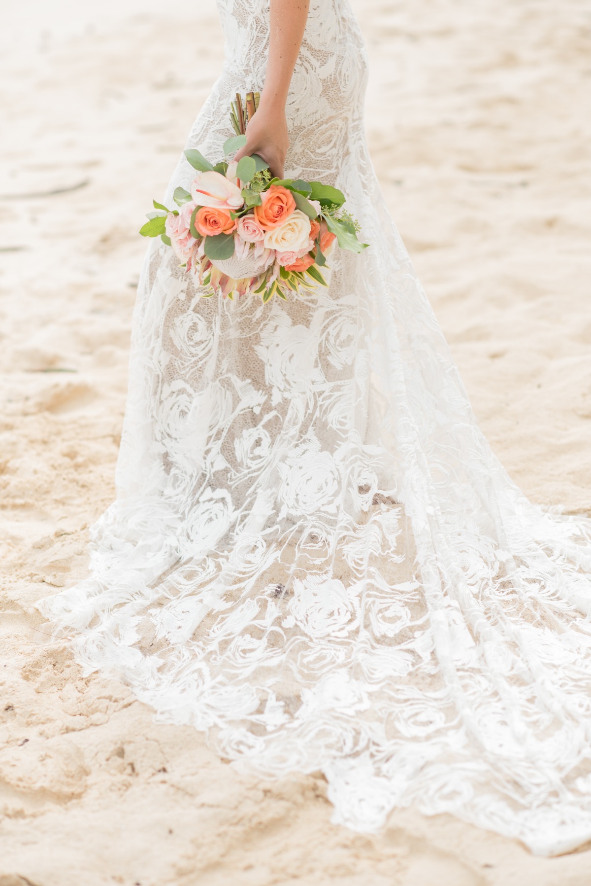 Bride holding a floral bouquet, wearing a lace wedding dress on a beach