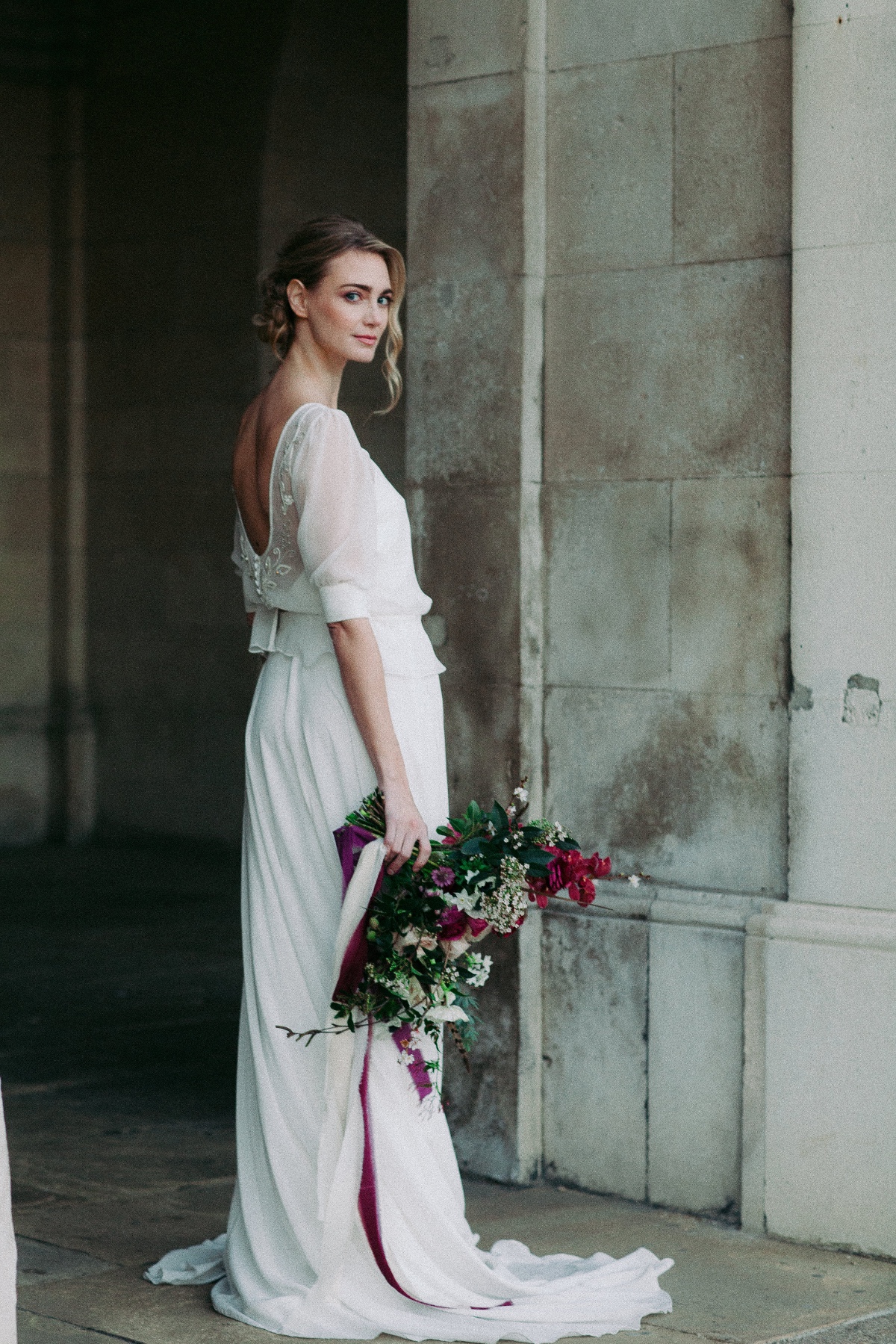Bride in flowing gown holds bouquet at stone archway