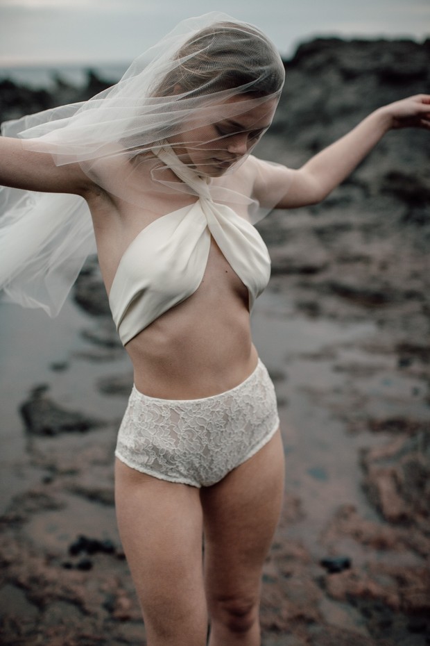 A woman in white lingerie and veil poses on a rocky beach