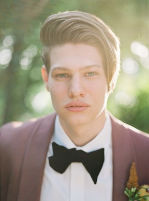 Groom with sleek hair wears burgundy suit and bow tie