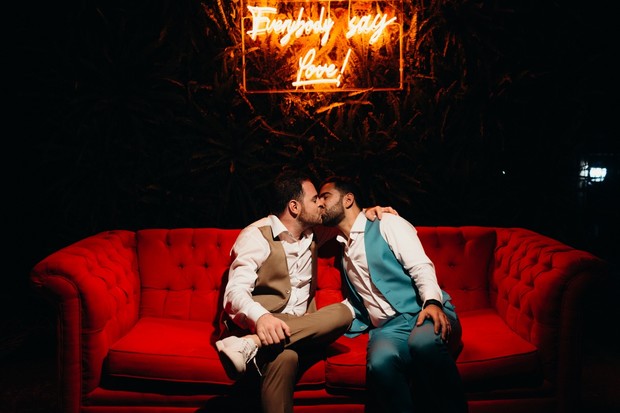 Gay couple kissing under neon love sign on red couch