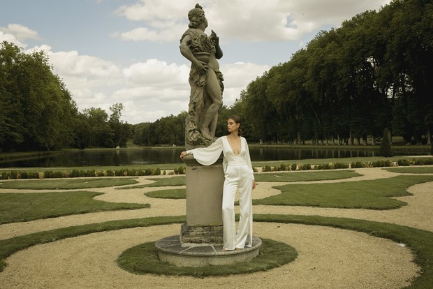 Woman in white jumpsuit poses by antique statue in formal garden