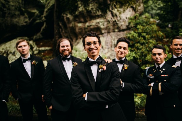 Groomsmen in black tuxedos smile for a wedding photo outdoors with trees