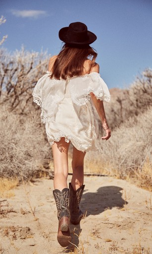 Woman in cowboy boots and dress walks away in desert