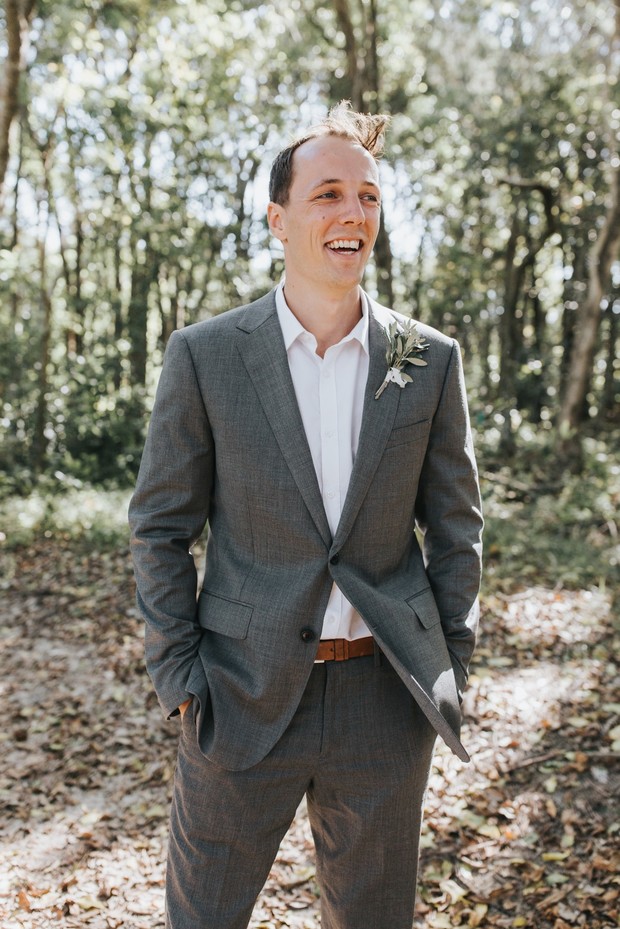 Smiling groom in grey suit stands in forest with hands in pockets