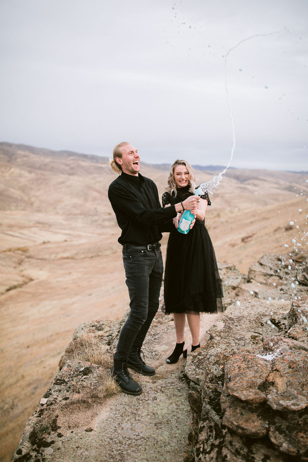 Excited couple pops champagne with celebratory spray on a cliff