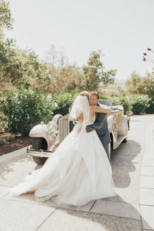 Bride and father share a wedding hug beside vintage car