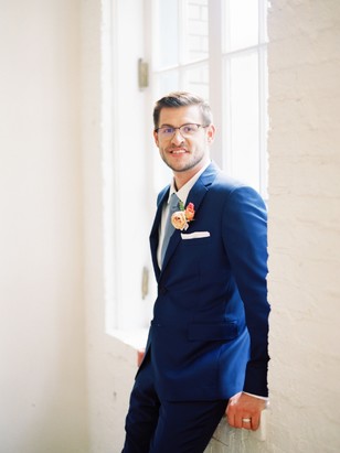 Groom in blue suit poses near window for wedding photos
