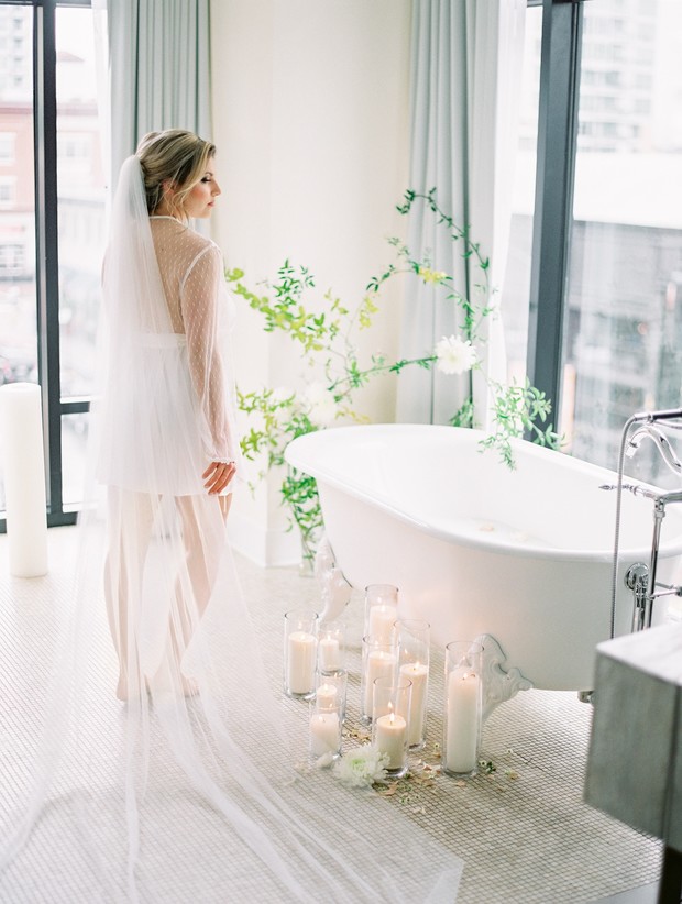 Bride in white robe and veil near bathtub with candles