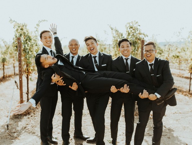 Groomsmen joyfully lifting a laughing friend in vineyard