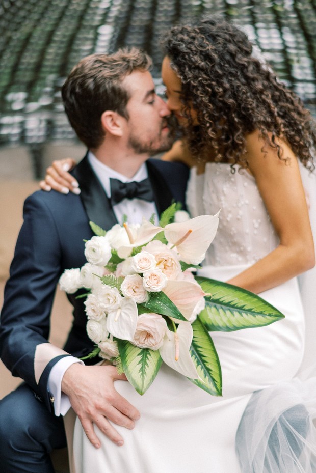 Newlyweds share a kiss while holding a beautiful floral bouquet
