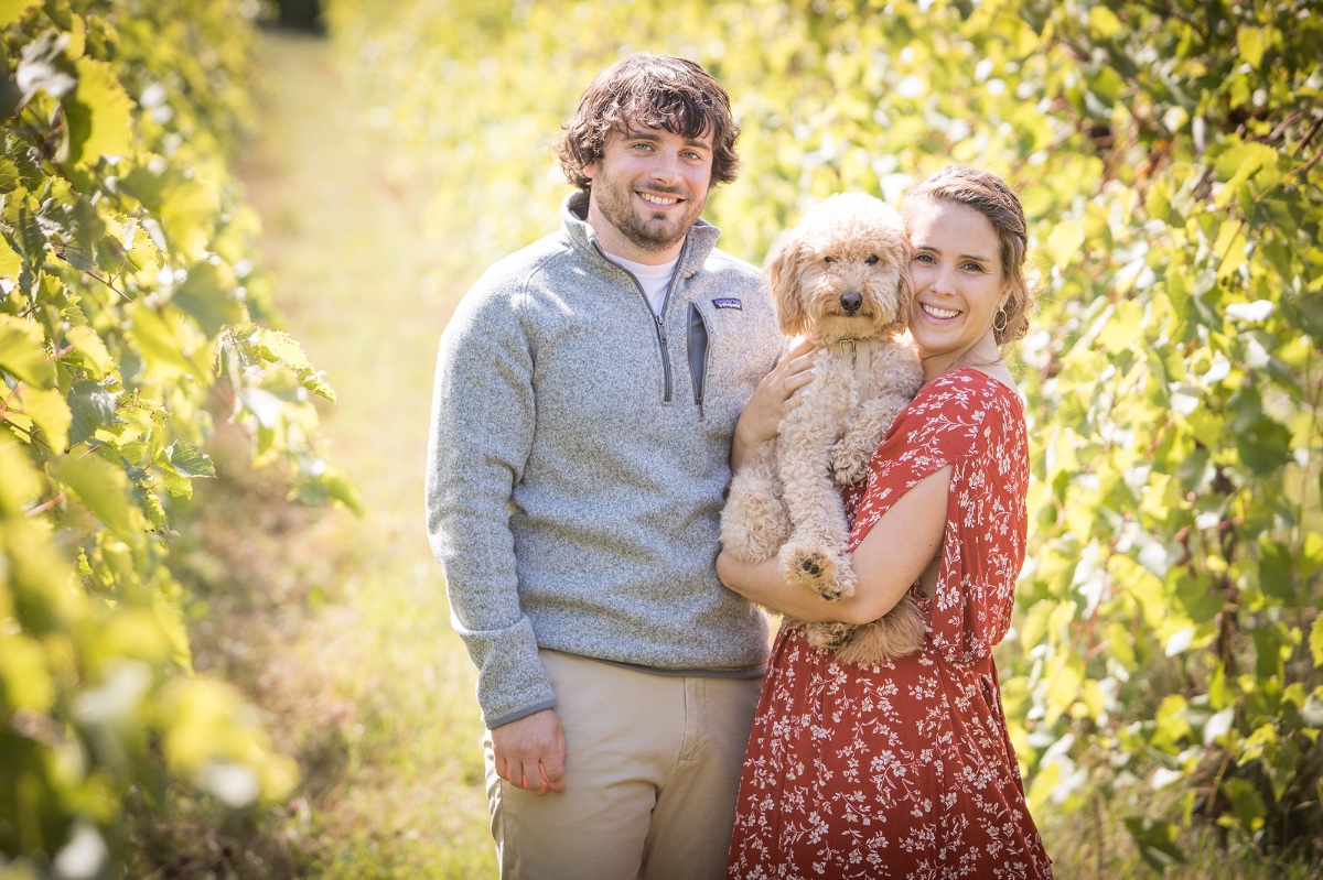 A happy couple with their dog in a vineyard smiles at the camera