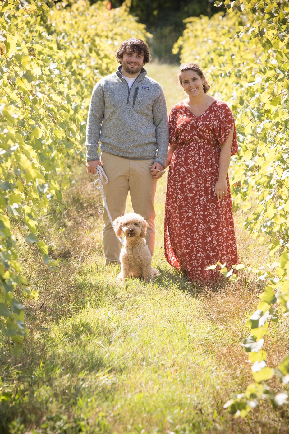 Couple with dog pose in vineyard, smiling at the camera