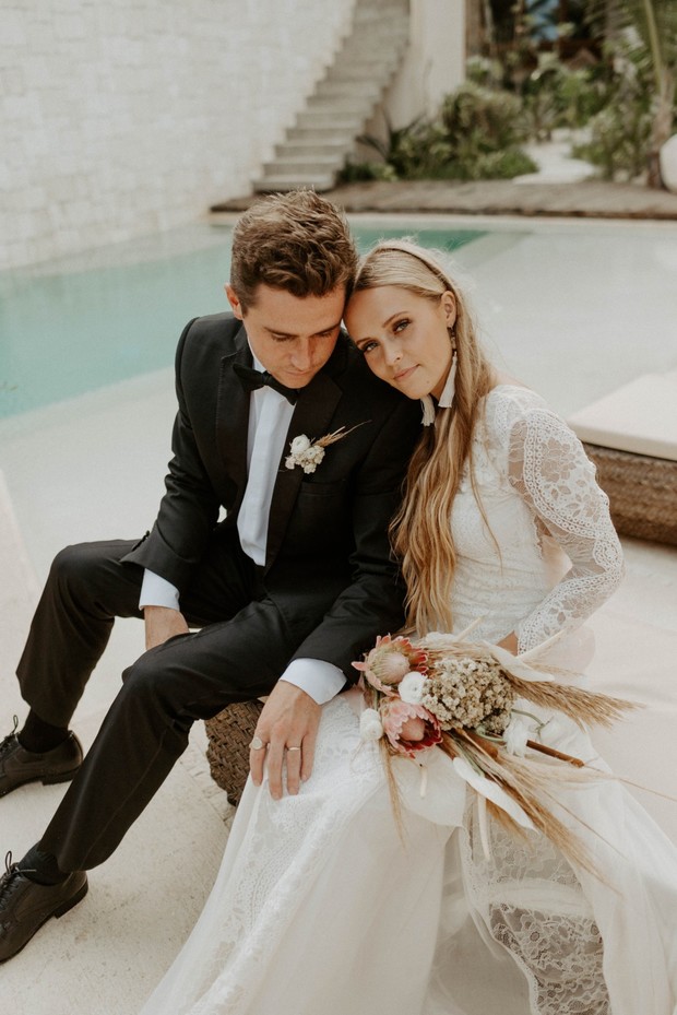 Newlyweds in formal attire sit by the pool on their wedding day