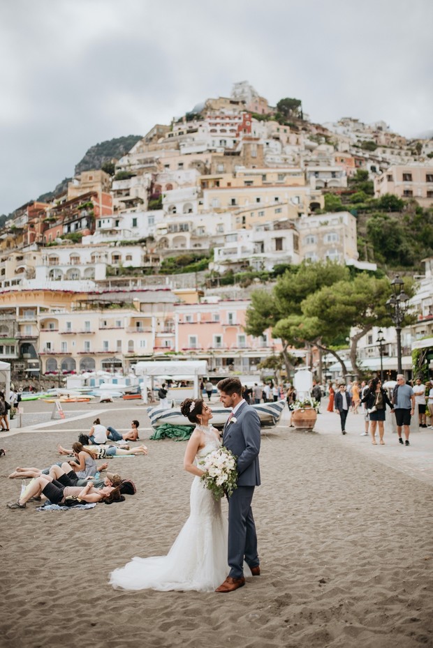 Bride and groom pose on Italian beach with village backdrop