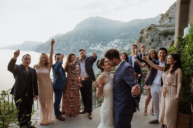 Newlyweds share a kiss with friends holding sparklers at wedding