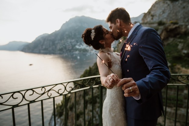 Newlyweds kiss with sparklers on a balcony overlooking the ocean