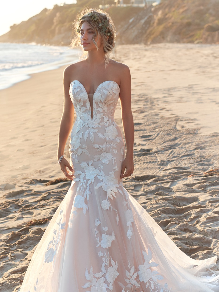 Bride in a floral wedding dress stands on a sunny beach
