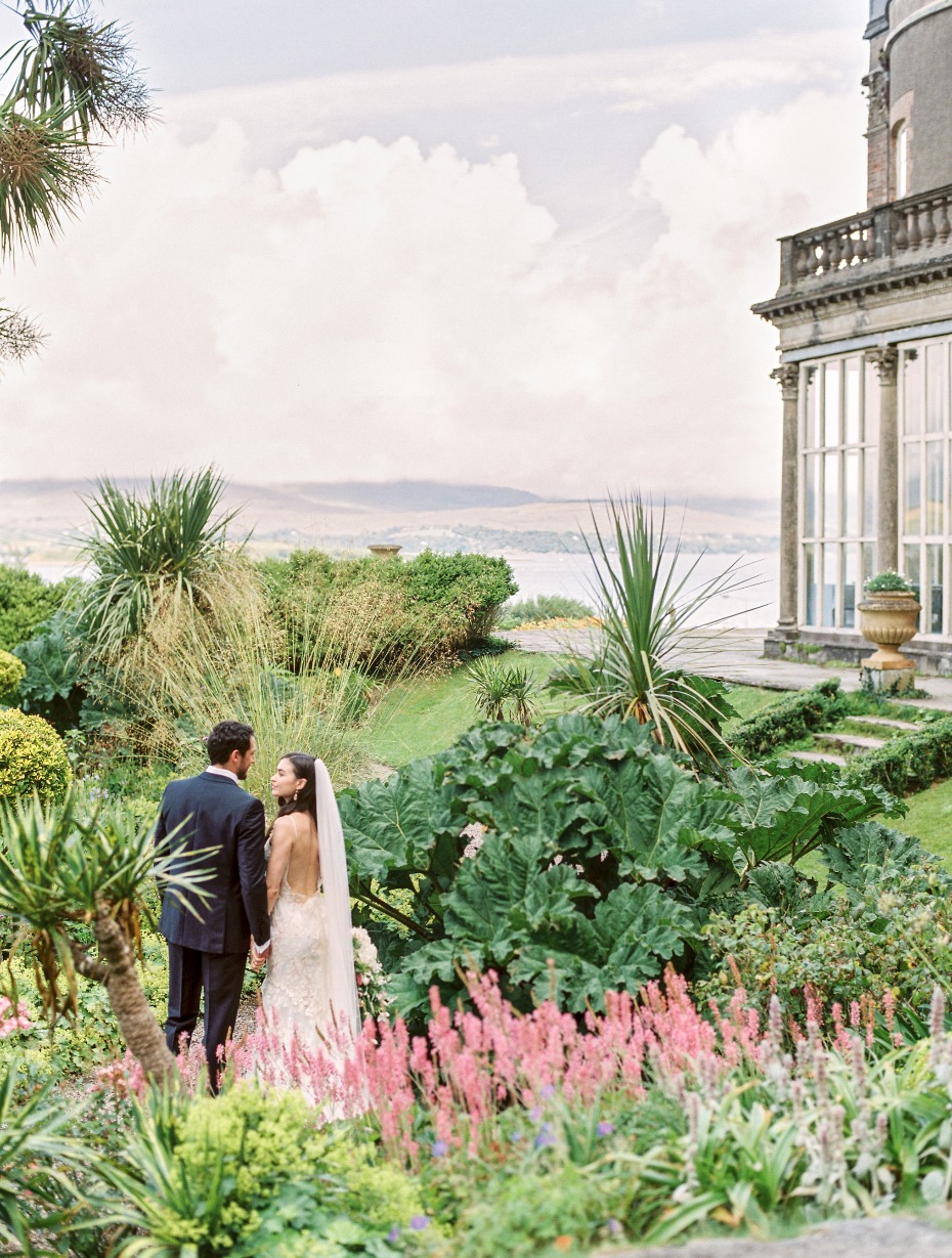 Romantic wedding couple strolls through a lush garden overlooking the sea