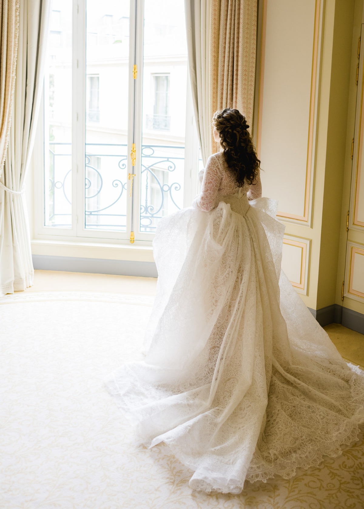 Bride in wedding dress looking out a window near curtains