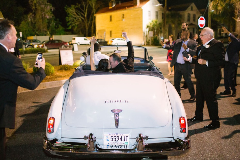 Newlyweds kiss in vintage Oldsmobile convertible, cheered on by guests