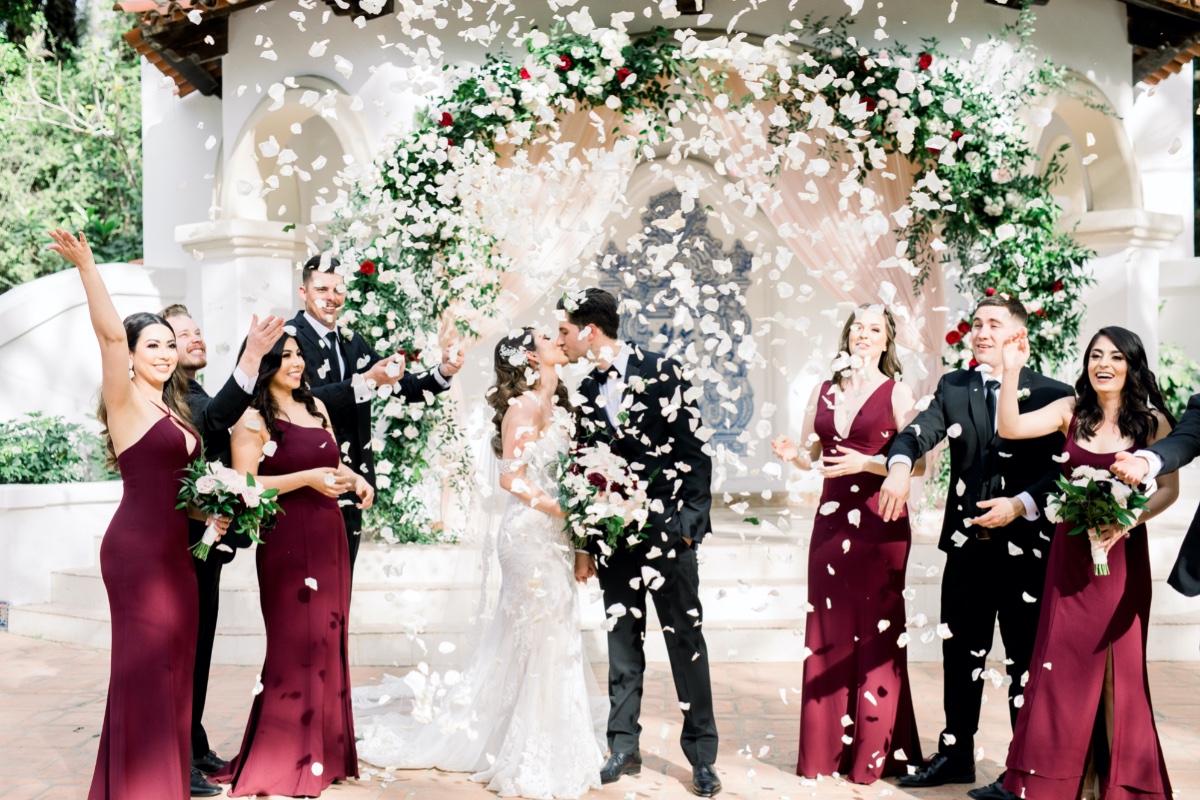 Newlyweds kiss amidst falling petals wedding party cheers