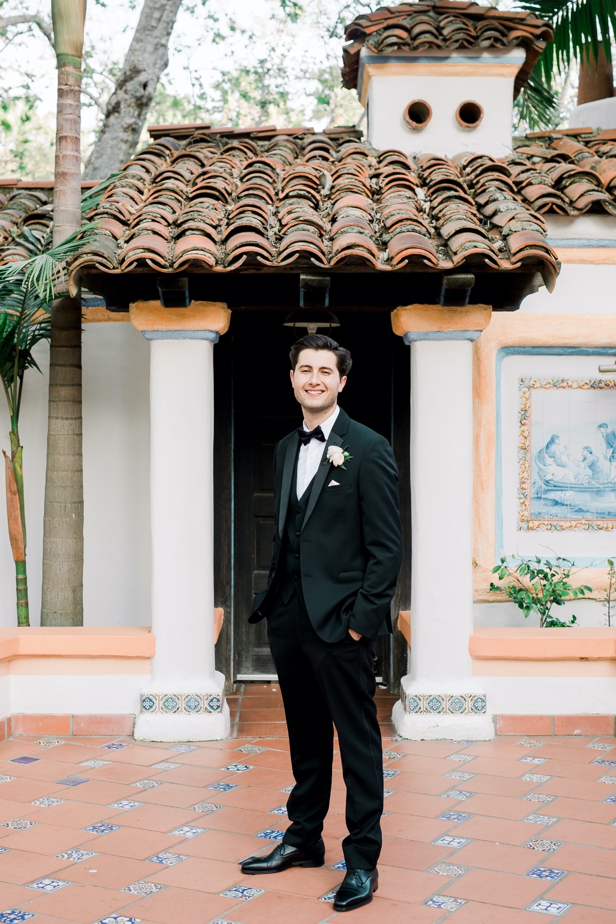 Groom in a black tuxedo poses near a tile roof building