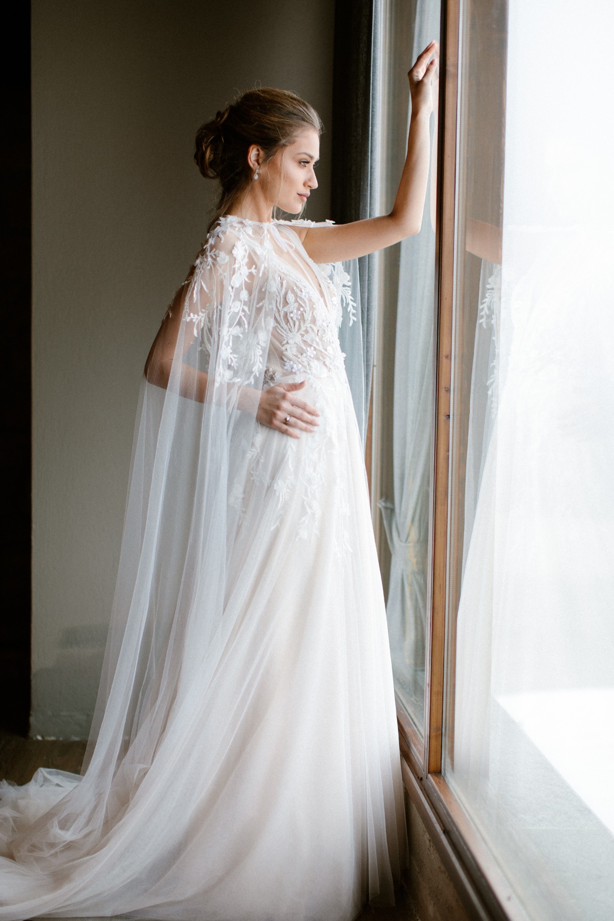 Serene bride in lace dress and cape gazes out window