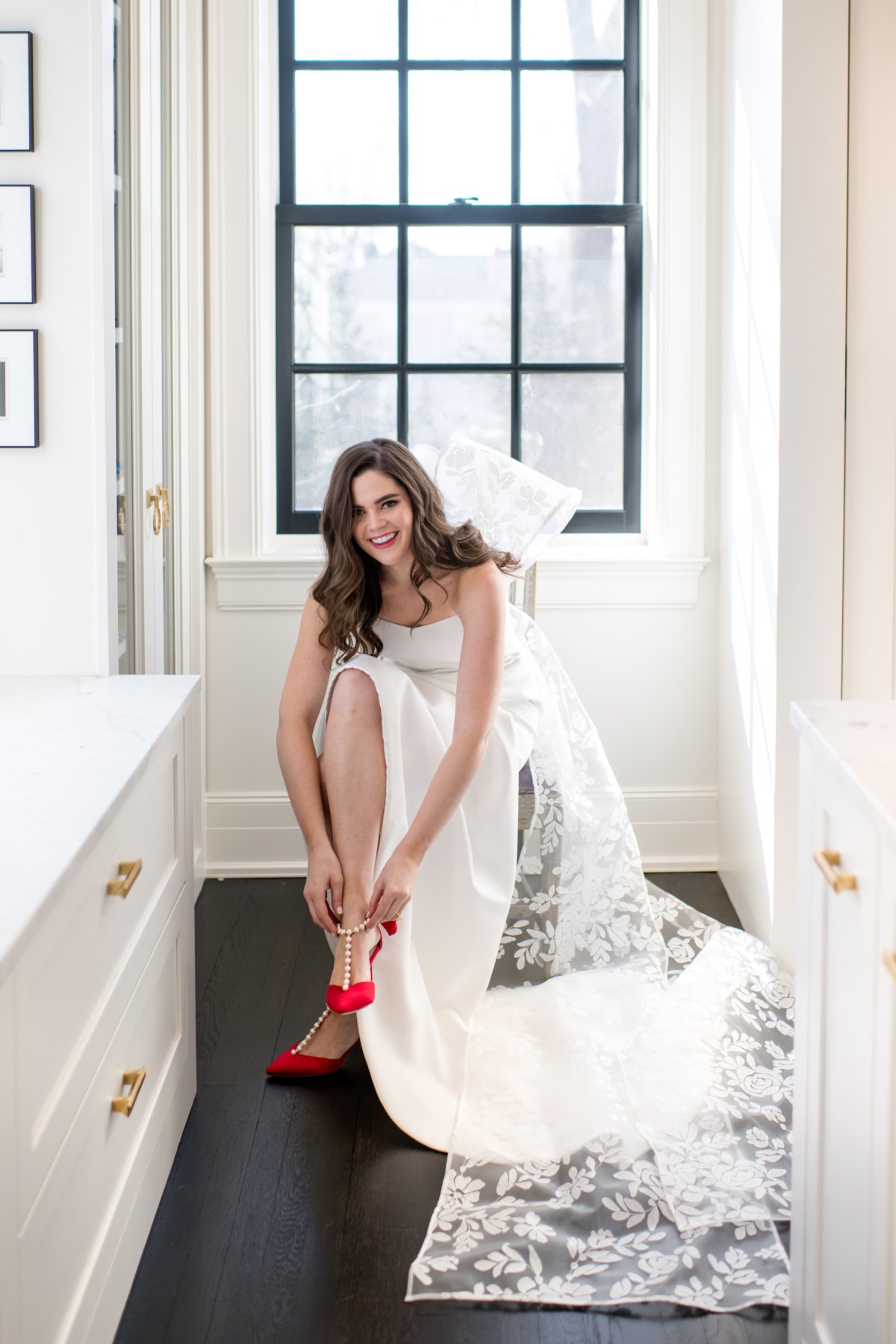 Bride smiles as she puts on her red wedding shoes in a dress