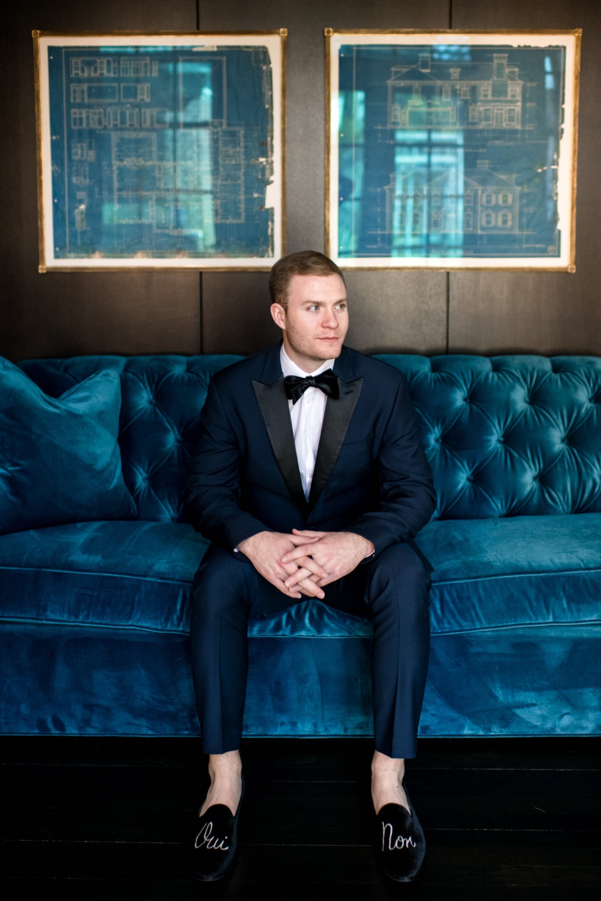 Groom in tuxedo sitting on a blue sofa with Oui Non slippers