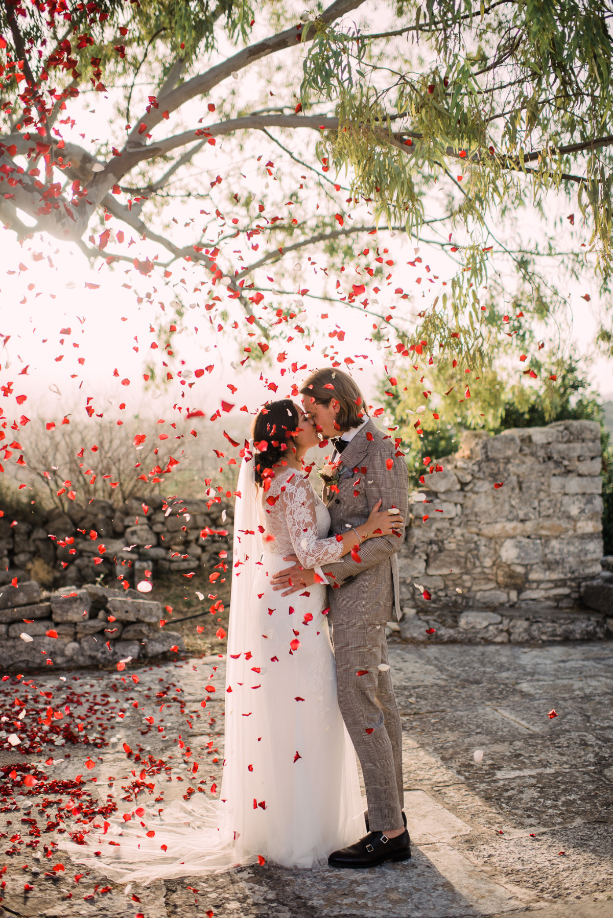 Bride and groom kiss under falling red petals at wedding