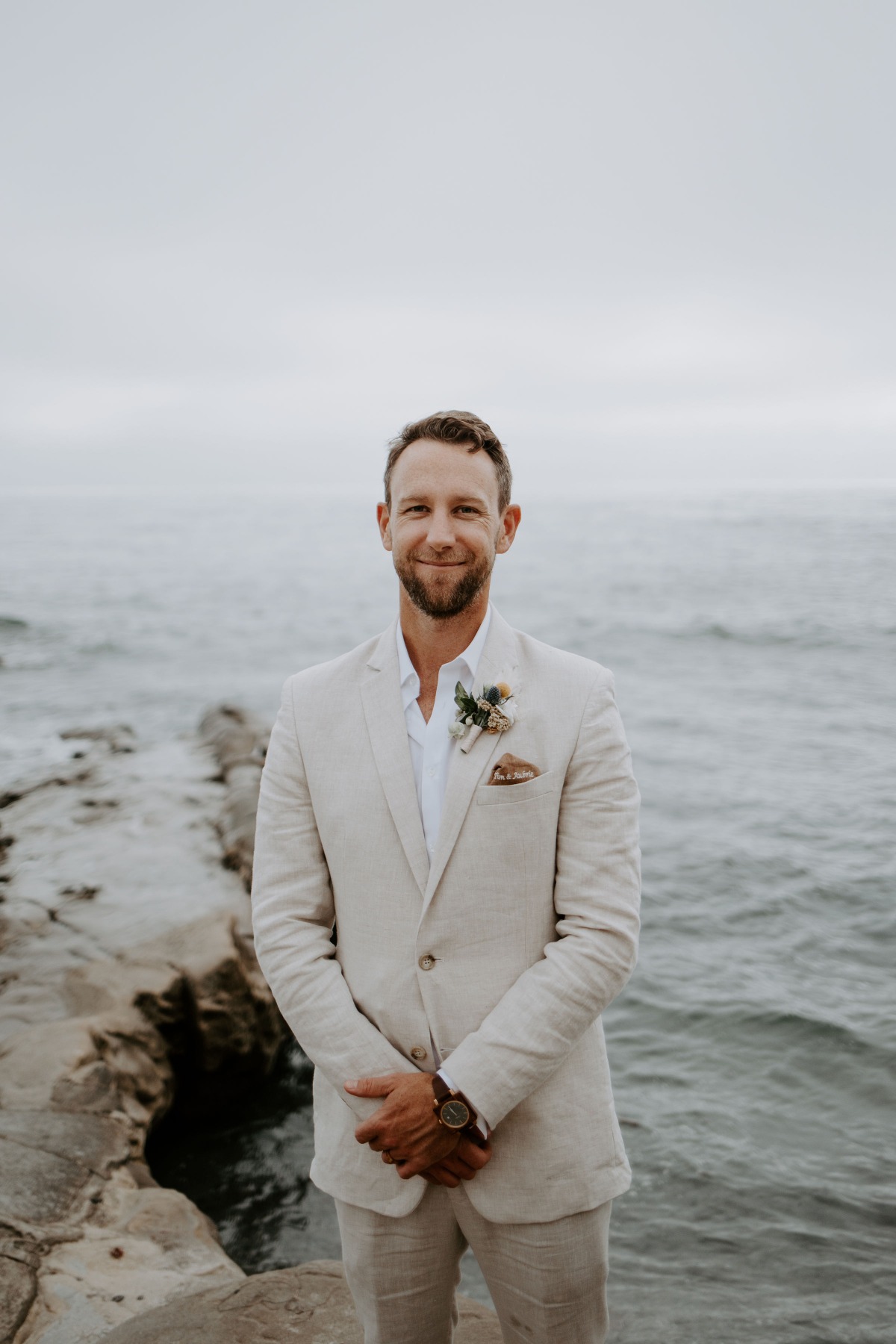 Smiling groom wearing a beige suit posing near the ocean