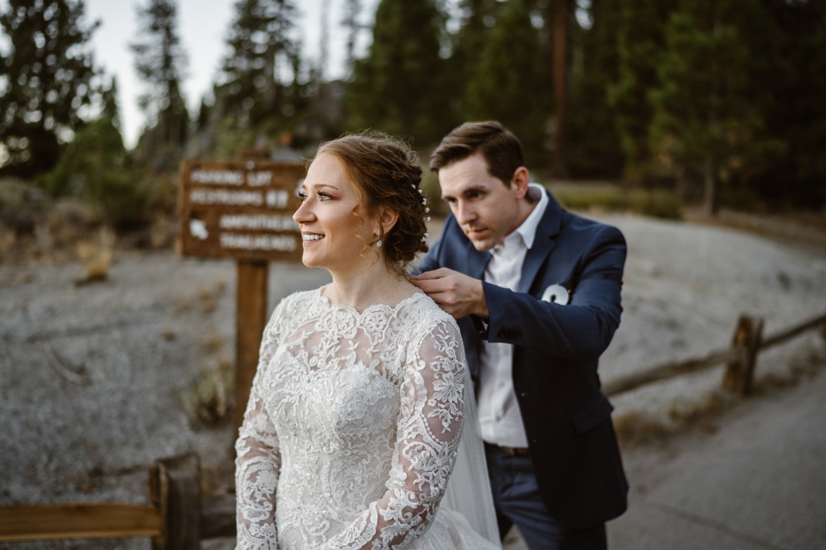 Groom adjusts smiling brides wedding dress outdoors