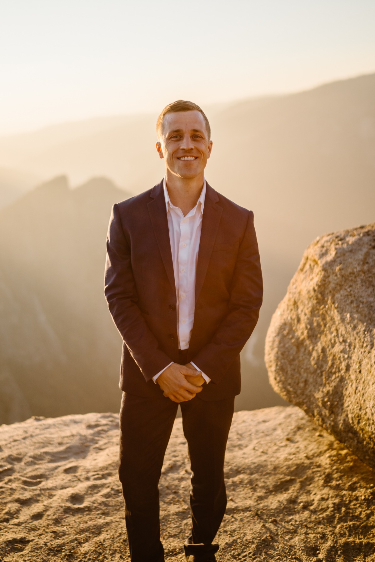 Man in a suit smiling on a mountain peak during sunset