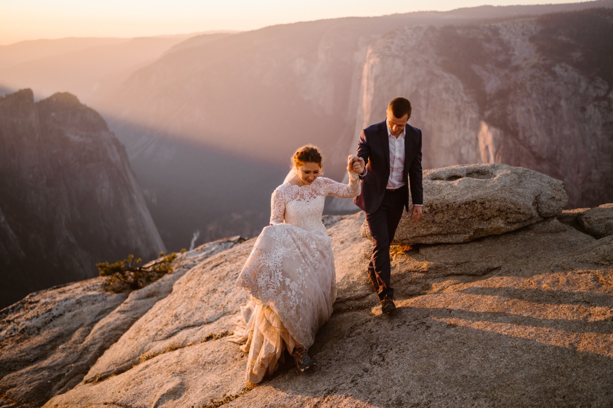 Newlyweds hike hand in hand on mountain rocks at sunset