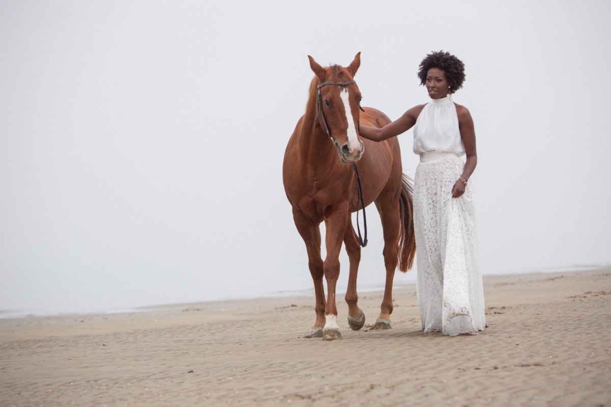Woman in white dress walks a brown horse on the beach