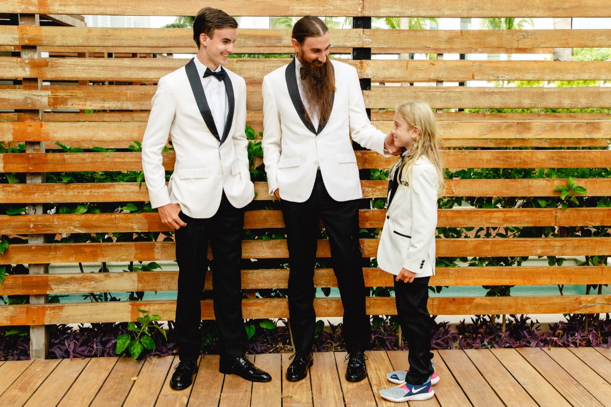 Three men in tuxedos smiling at a tropical wedding