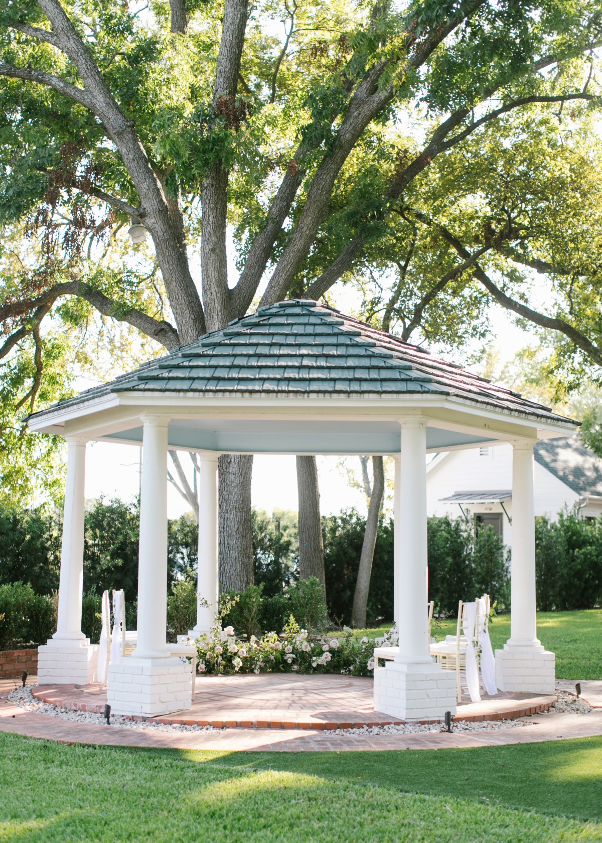 White gazebo beneath a large tree in a sunny wedding setting