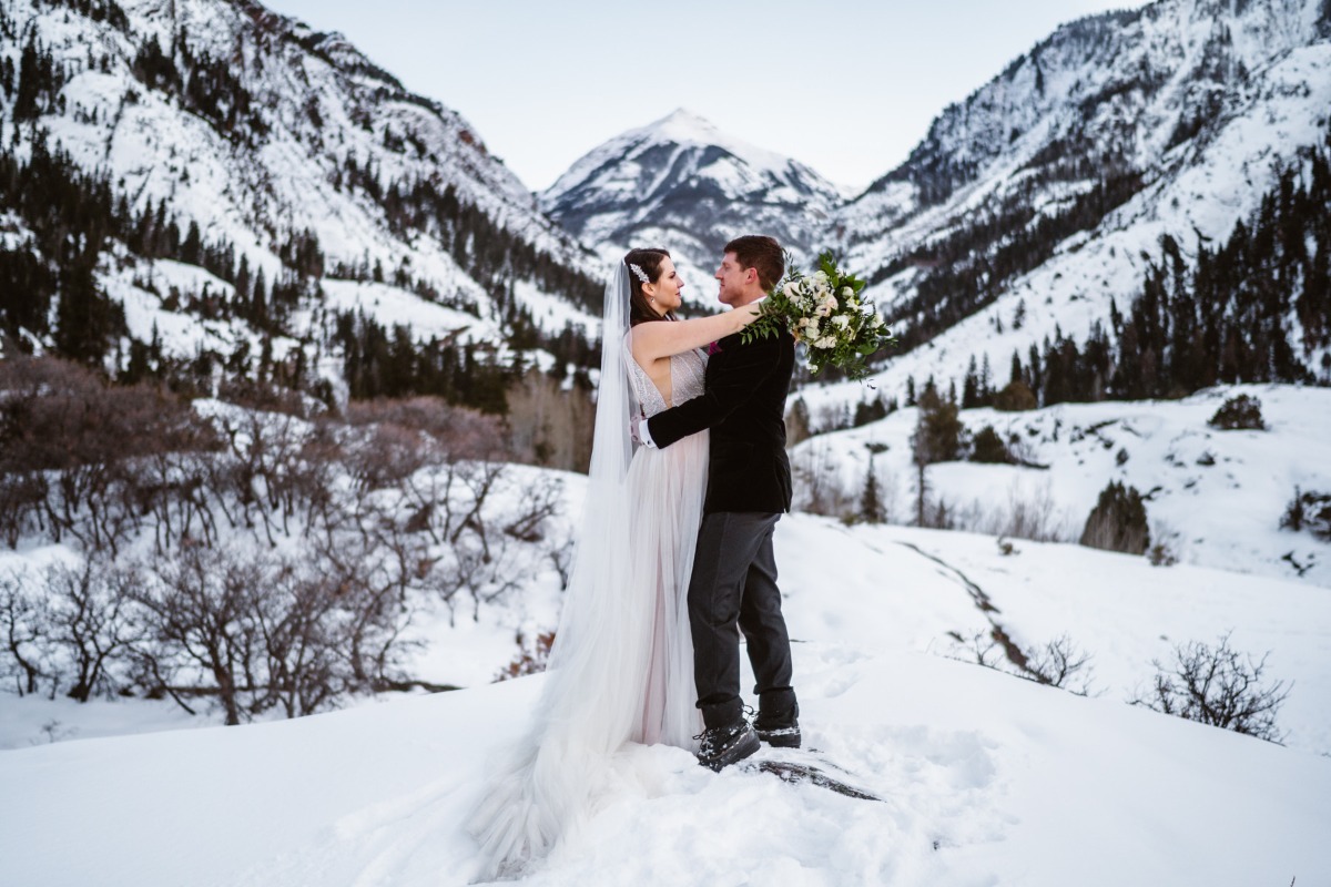 Winter wedding couple embraces with snowy mountain backdrop