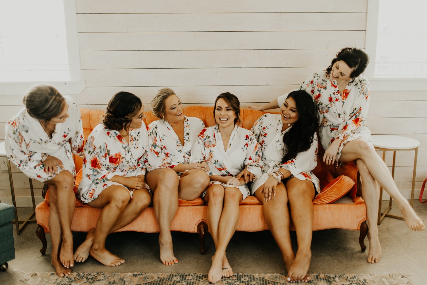 Bridesmaids in floral robes laughing on an orange sofa, pre-wedding fun