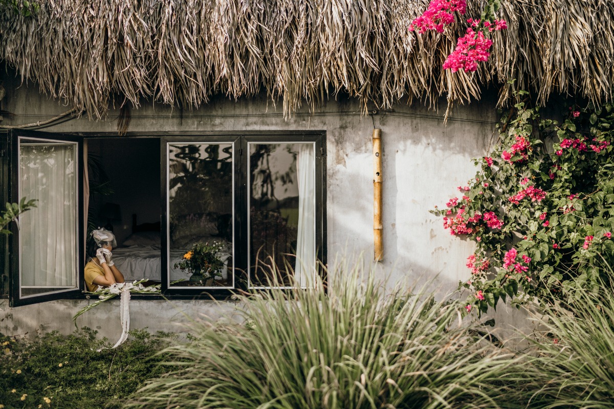Woman looks out window of a flowered thatched roof house