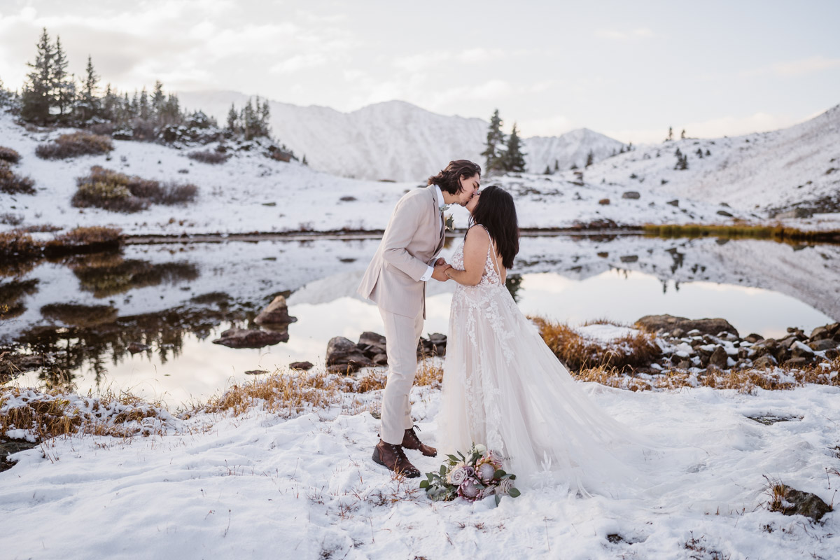 Newlyweds share a kiss by a snow-covered lake with mountain views