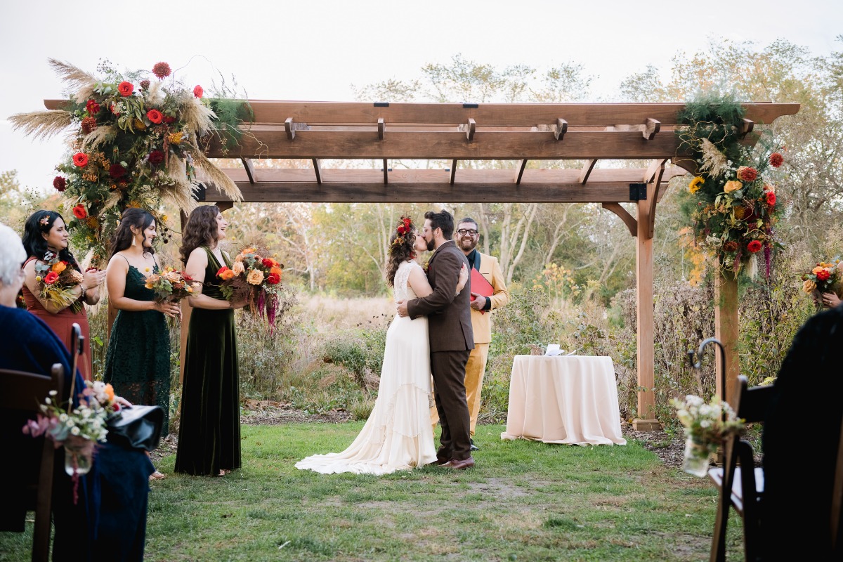 Newlyweds share a kiss under a wooden wedding arbor during ceremony