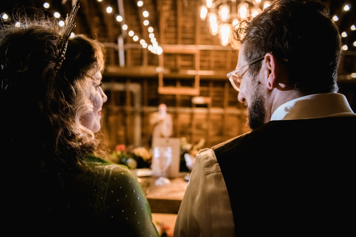 Couple in a rustic barn, softly lit, looking at the ceremony
