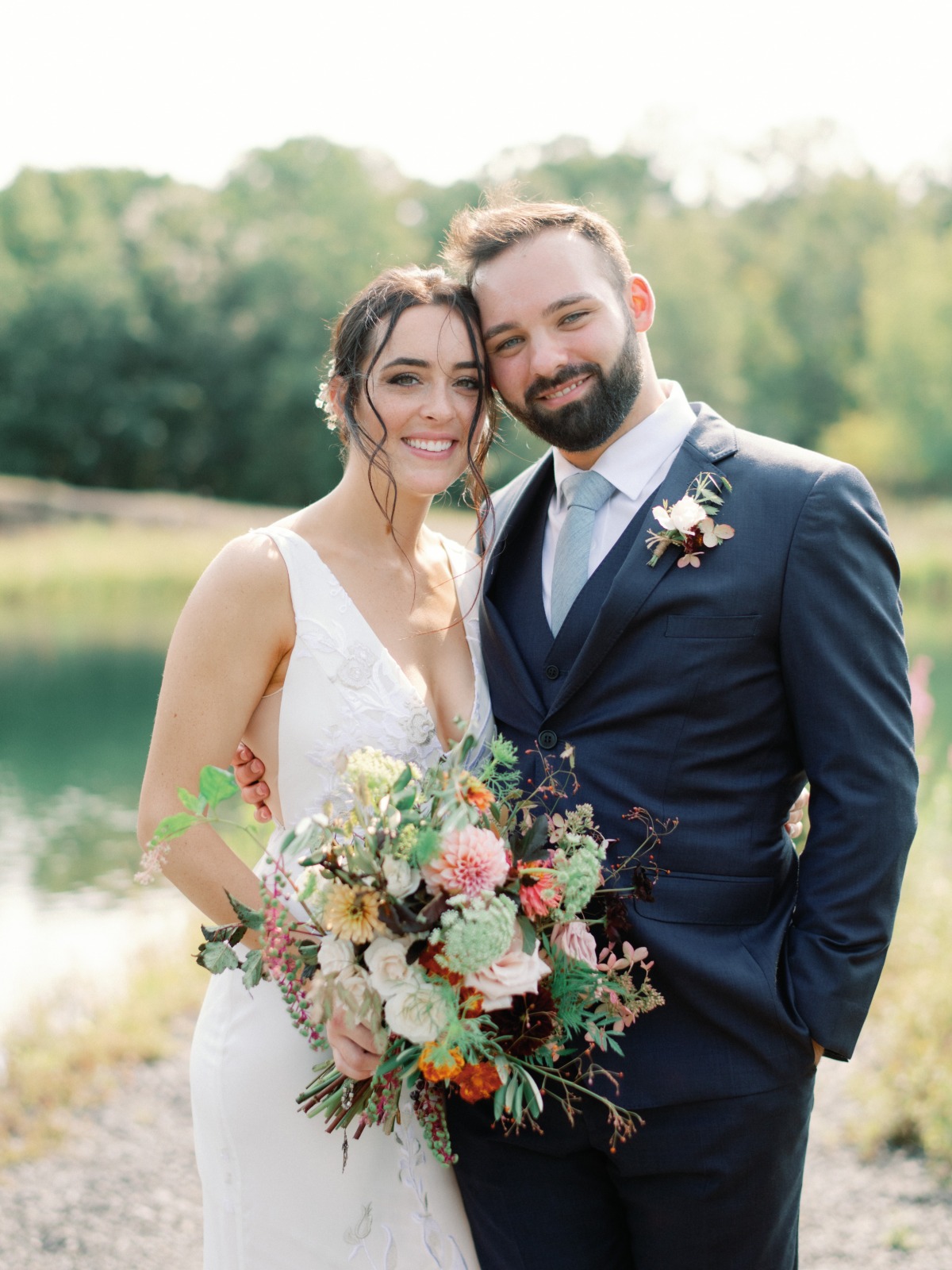 Smiling newlyweds pose by a lake with a colorful bouquet