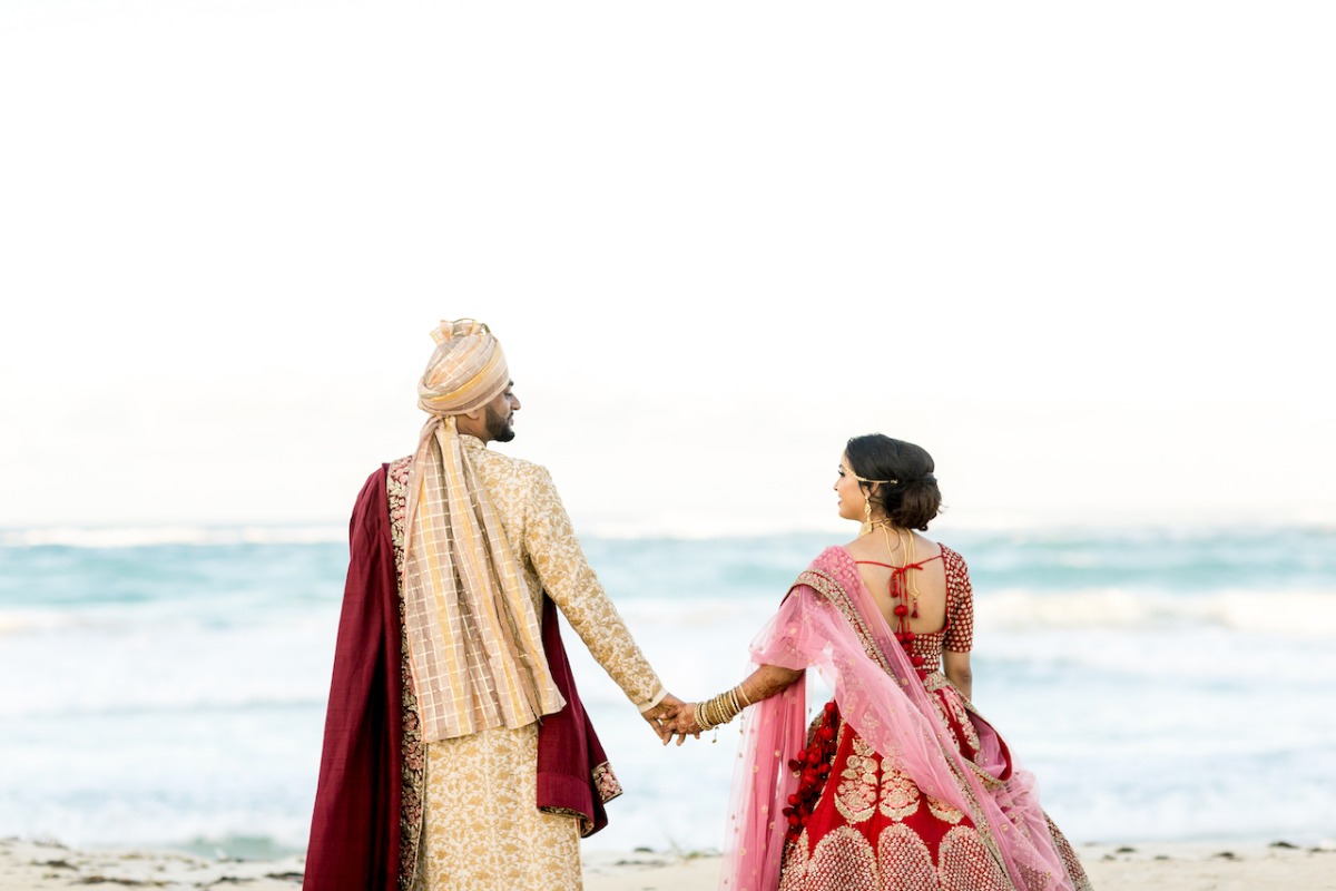 Bride and groom in traditional Hindu attire facing towards water on beach