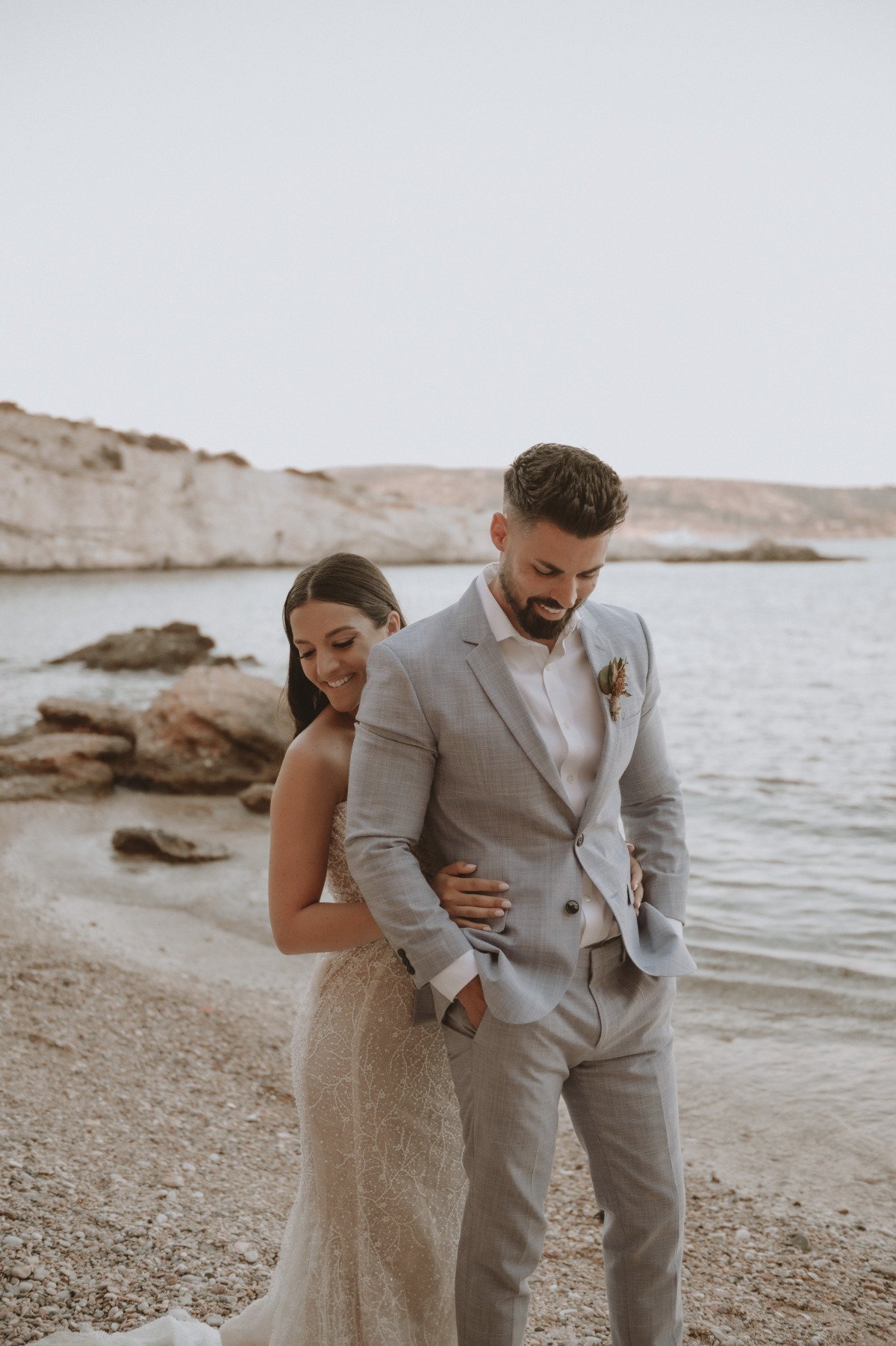 Bride hugging groom from behind on beach at first look
