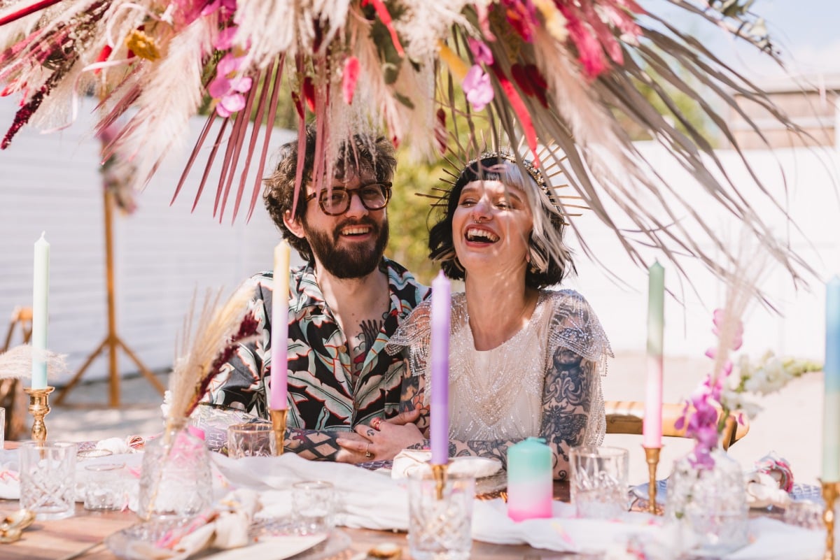 Bride and groom seated at reception table under floral installation laughing