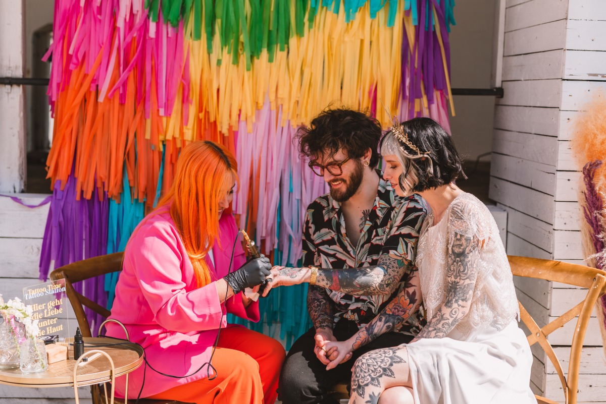 Couple receiving wedding tattoos in front of rainbow wall