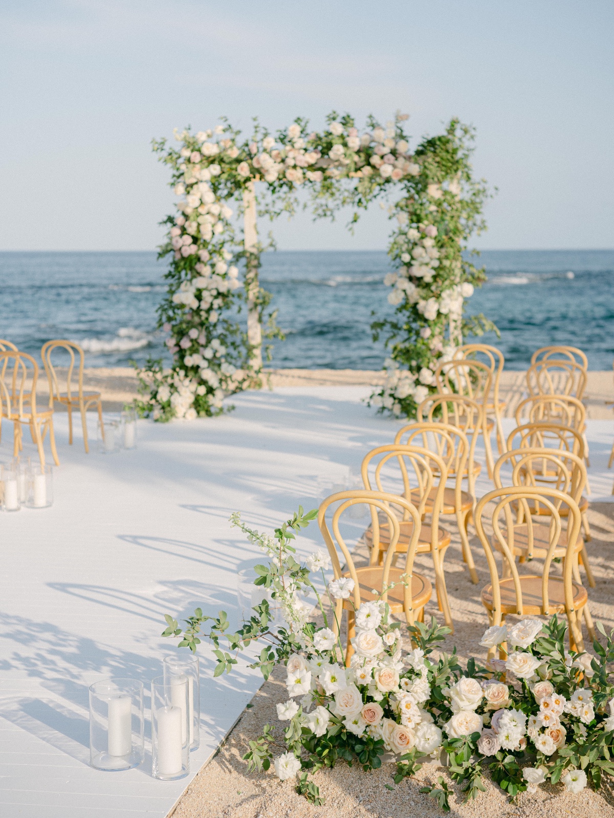 Ceremony area with floral arch, rattan chairs, and white roses floral arrangements next to aisle entrance